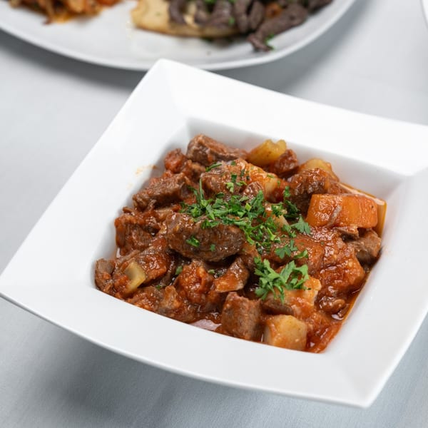 A white square bowl filled with a rich, red stew of beef and potatoes, garnished with parsley.