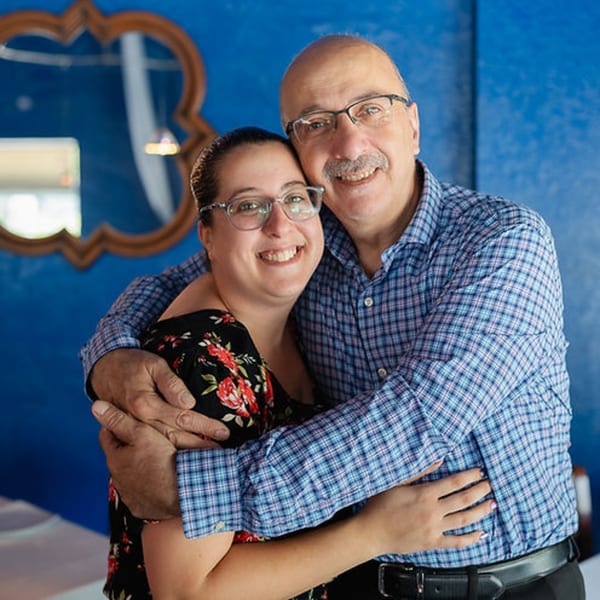 Smiling man and woman, possibly owners, embracing in front of a vibrant blue wall at Leila Restaurant.