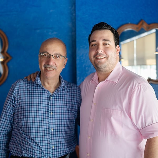 Two men, one older with glasses and a mustache, the other younger, smiling against a blue wall.